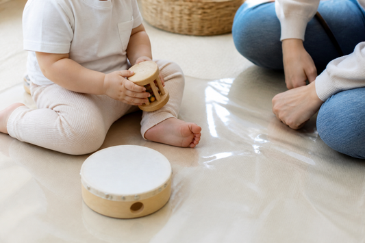 A young child exploring a shaker and small drum with a supervising adult in a clear home play area